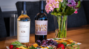bottles of clay shannon and ovis wines on a table with a vase of flowers and a colorful vegetable platter